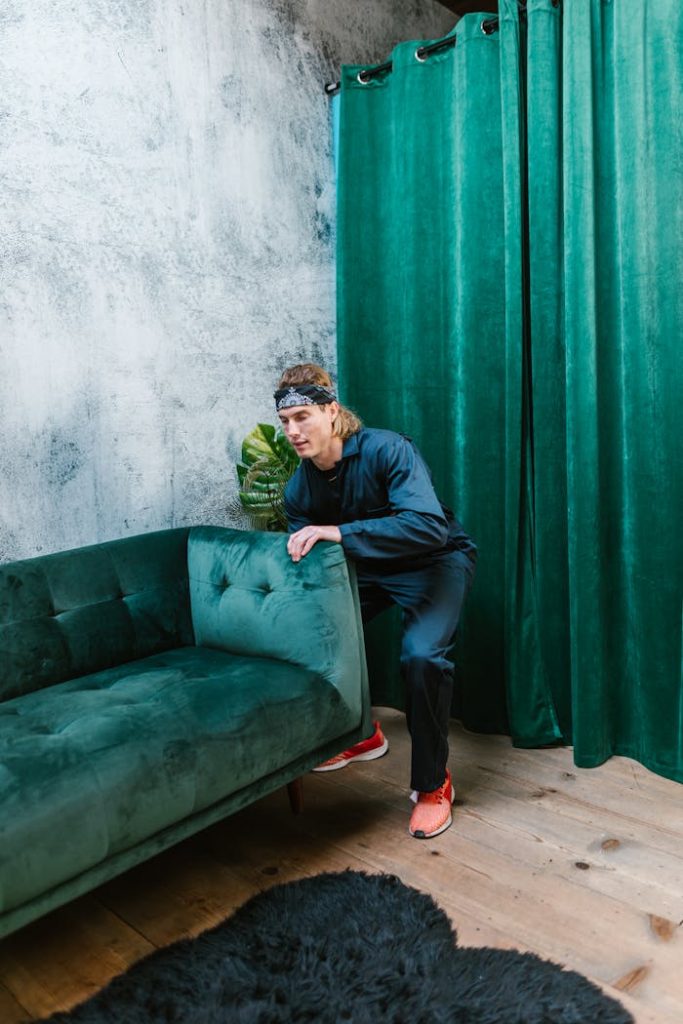 A man carefully moves a lush green velvet sofa in a modern interior space with green curtains.